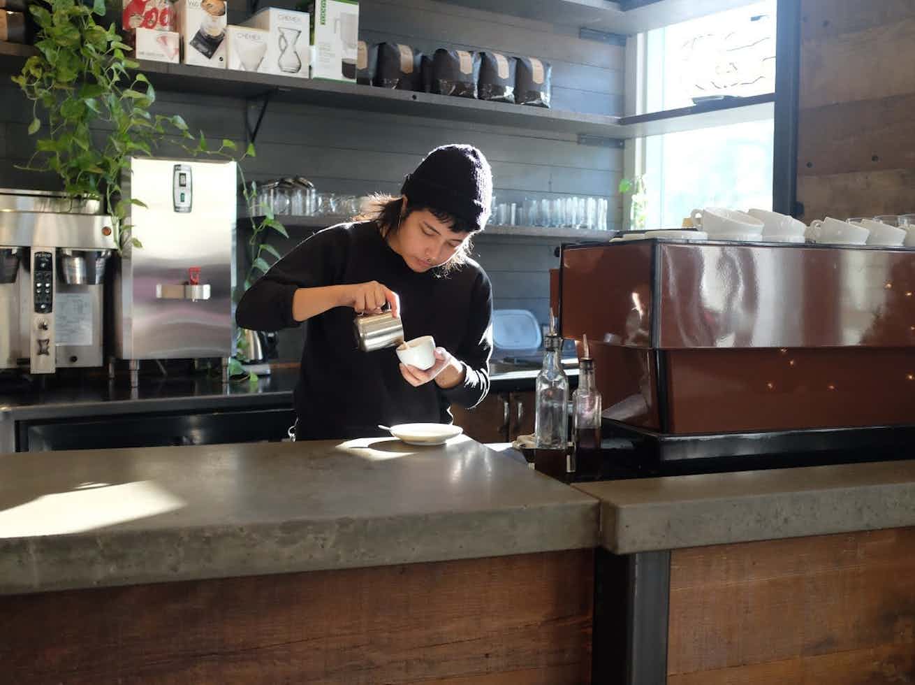 Barista pouring milk into a cup behind a café counter with a brown espresso machine and sunlit shelves.