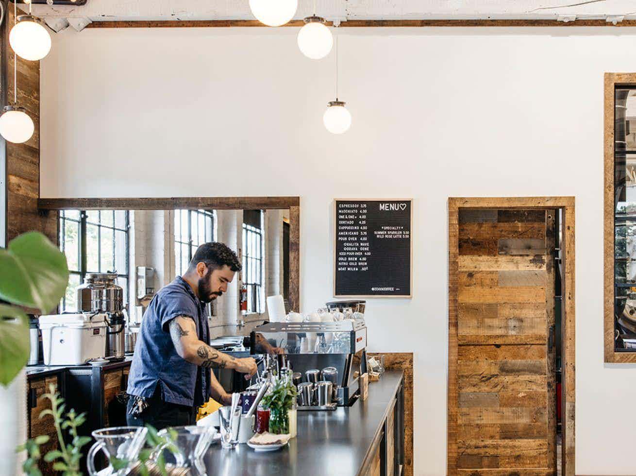  Open café interior with high ceilings, globe lights, and two baristas working behind a long counter beneath tall windows.