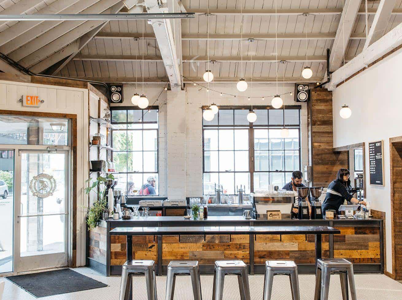  Barista preparing drinks at a modern café counter with wood accents, globe lights, and a wall-mounted menu.