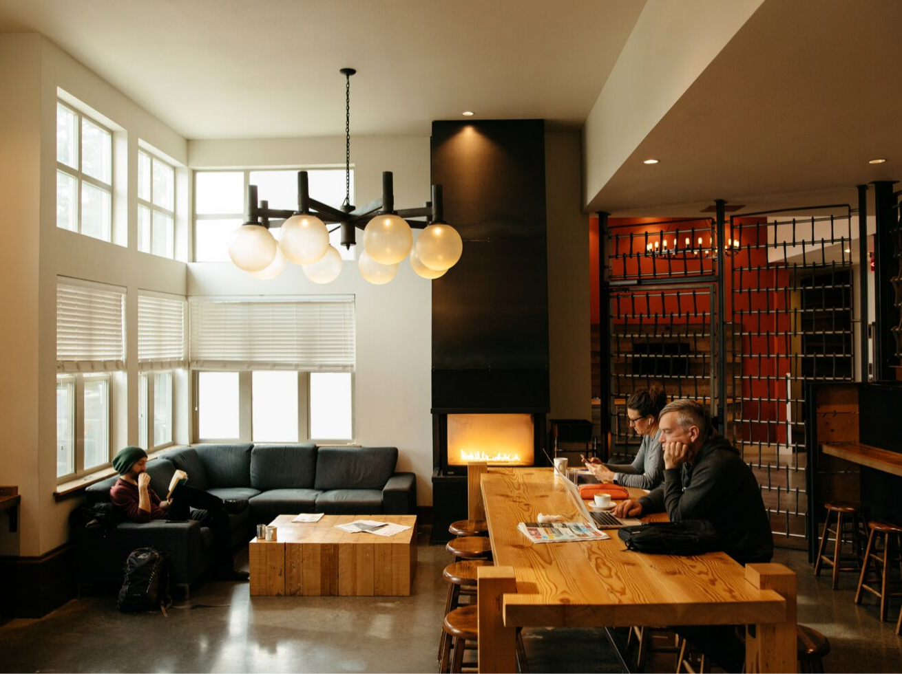 Cozy café interior with a modern fireplace, lounge seating, long communal table, and guests reading and working.